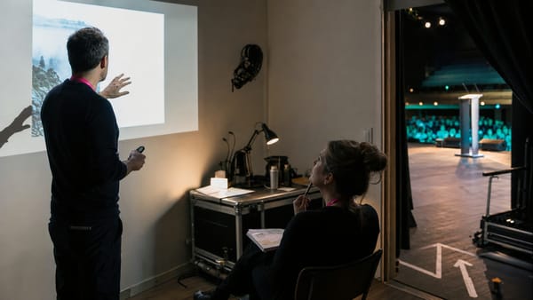 Un especialista ensaya una presentación con una productora en backstage mientras se observa un escenario al fondo.
